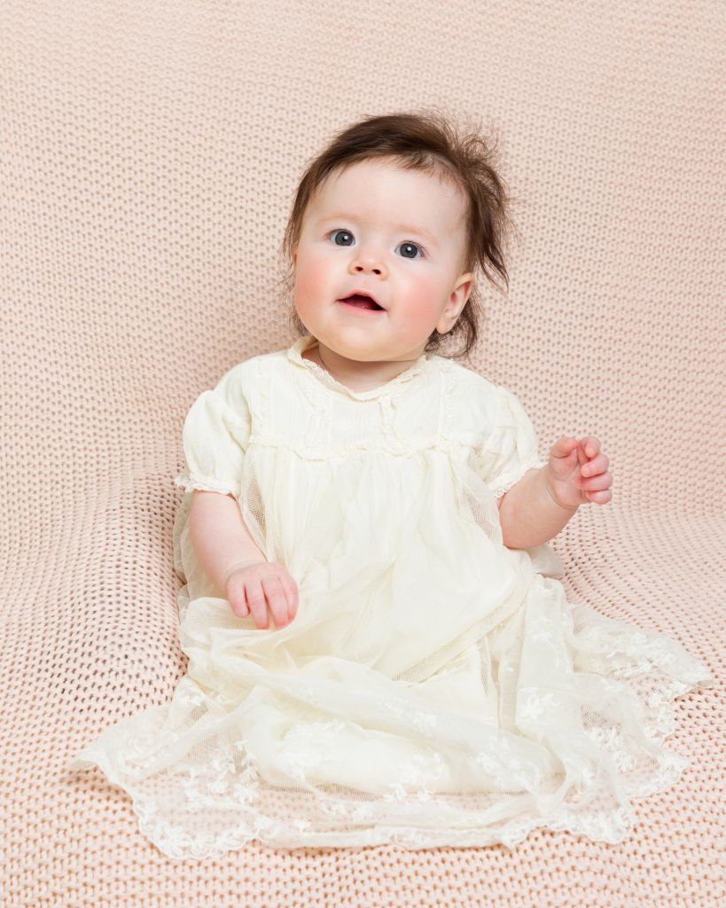 a little girl sitting propped on a beanbag in a christening dress