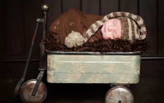 A newborn boy is swaddled in brown with a long tail hat and sleeping in a vintage styled trolley