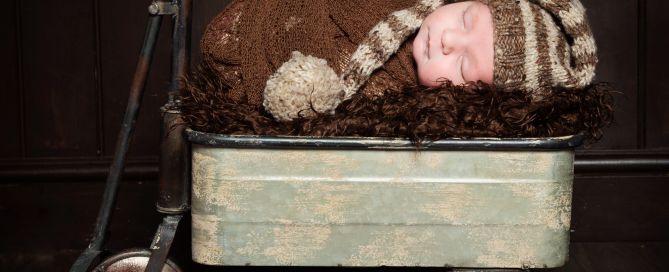A newborn boy is swaddled in brown with a long tail hat and sleeping in a vintage styled trolley