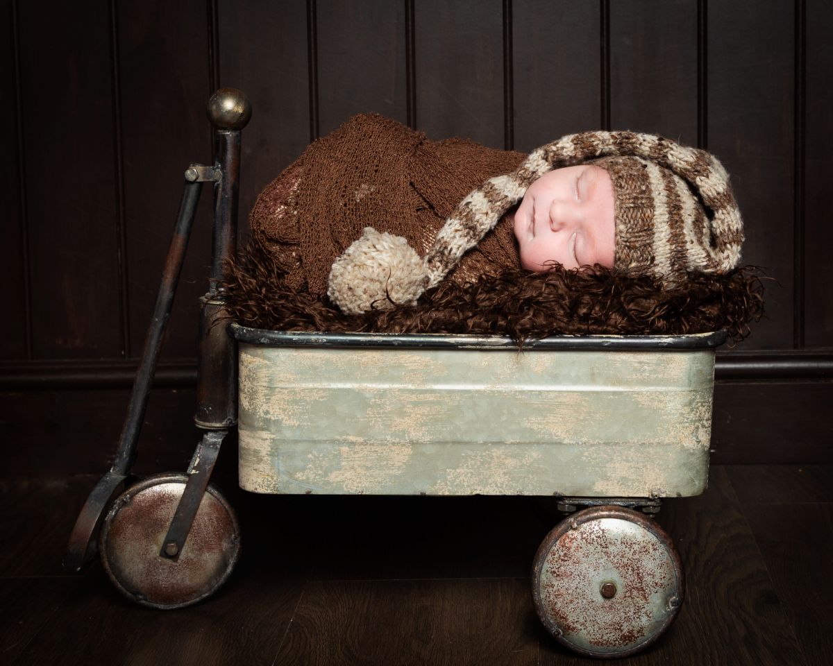 A newborn boy is swaddled in brown with a long tail hat and sleeping in a vintage styled trolley