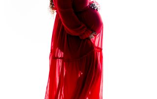 A woman poses in a flowing red maternity dress lit in silhouette