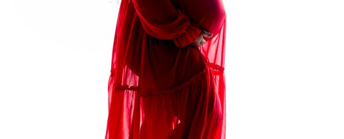 A woman poses in a flowing red maternity dress lit in silhouette