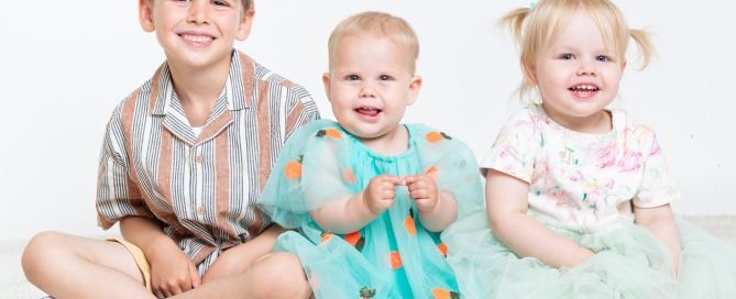 A big brother poses for a portrait photo with his two little sisters and they're all smiling.