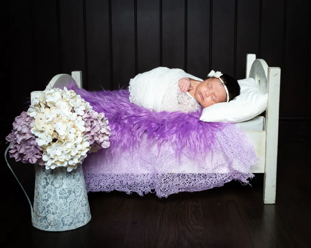 Newborn Photography East Grinstead West Sussex A newborn wrapped in white lies on a bed covered with purple fur and a vase of hydrangeas nearby