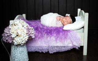 A newborn wrapped in white lies on a bed covered with purple fur and a vase of hydrangeas nearby