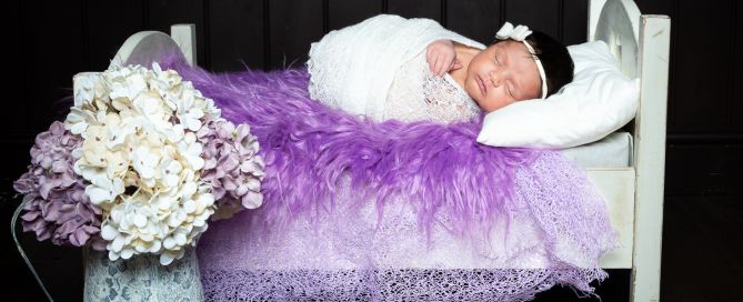 A newborn wrapped in white lies on a bed covered with purple fur and a vase of hydrangeas nearby