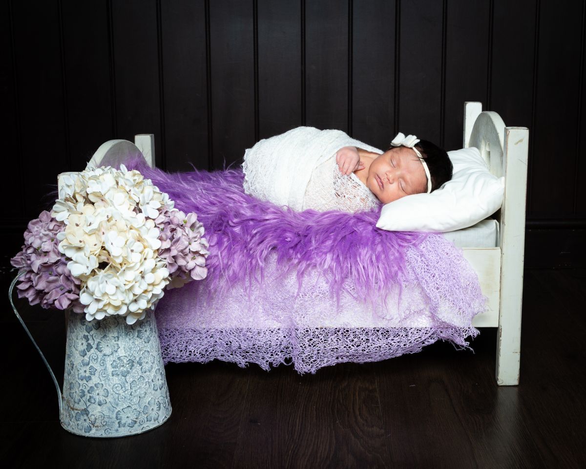 A newborn wrapped in white lies on a bed covered with purple fur and a vase of hydrangeas nearby