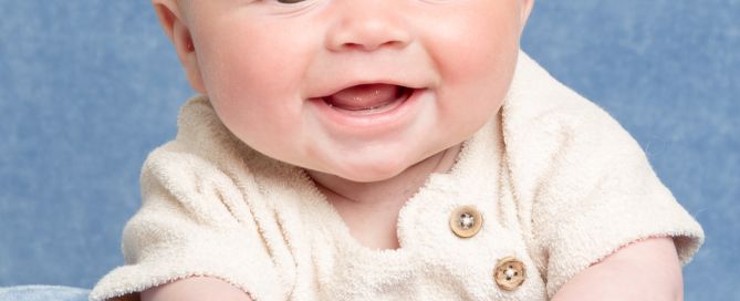 A smiling baby boy in a cream romper on a blue blanket