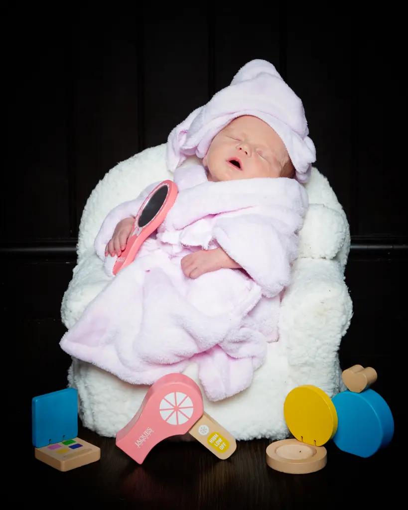 A sleeping newborn wearsa a pink bathrobe and a towel on her head and is relaxing on a spa chair