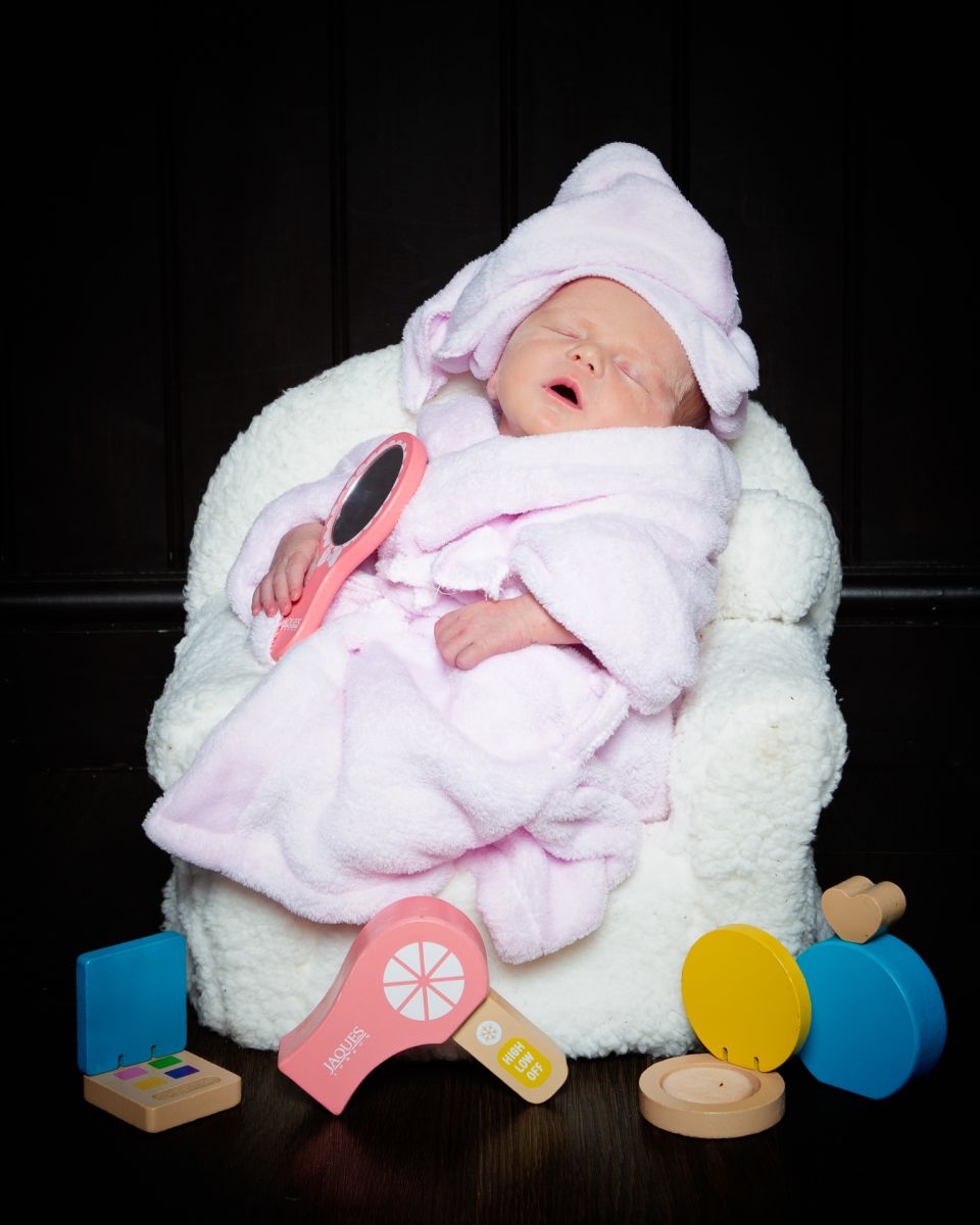 A sleeping newborn wearsa a pink bathrobe and a towel on her head and is relaxing on a spa chair