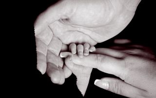 A newborn holding onto his mums hand with his dad's hand wrapped around them both in black and white