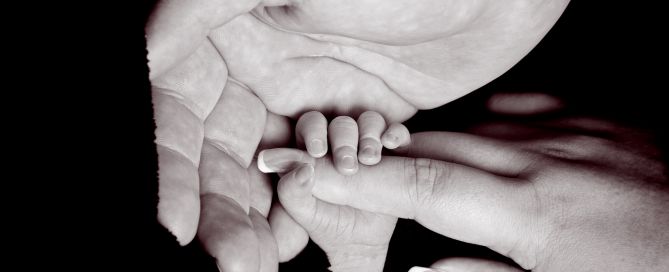 A newborn holding onto his mums hand with his dad's hand wrapped around them both in black and white