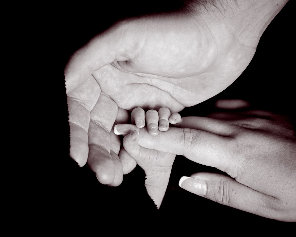 A newborn holding onto his mums hand with his dad's hand wrapped around them both in black and white