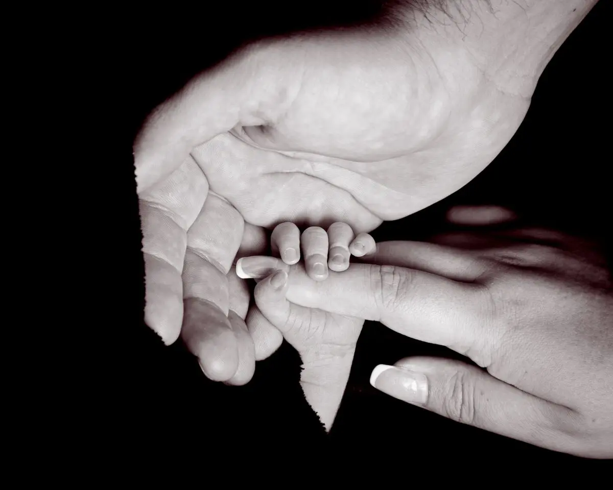 A newborn holding onto his mums hand with his dad's hand wrapped around them both in black and white