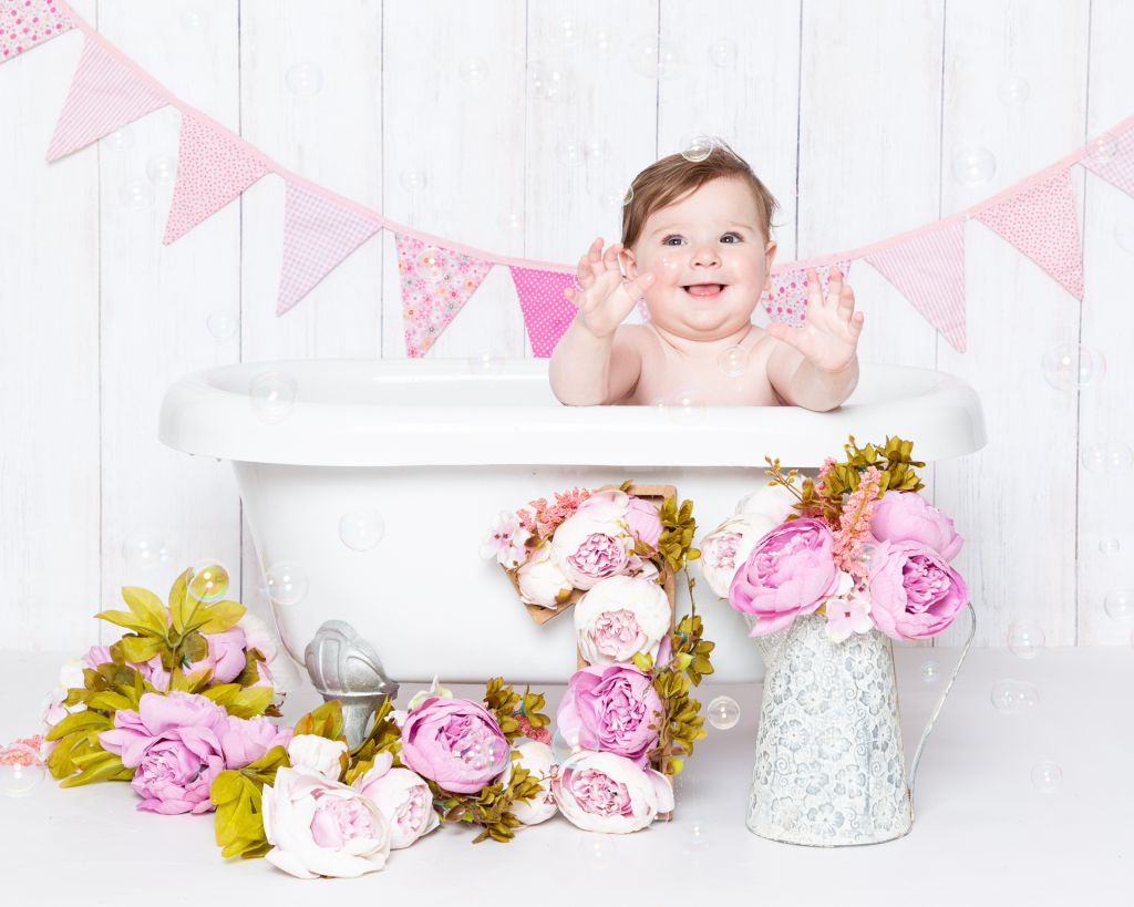 A little girl is excited to be in a bath tub against a white plank backdrop with floral accesories