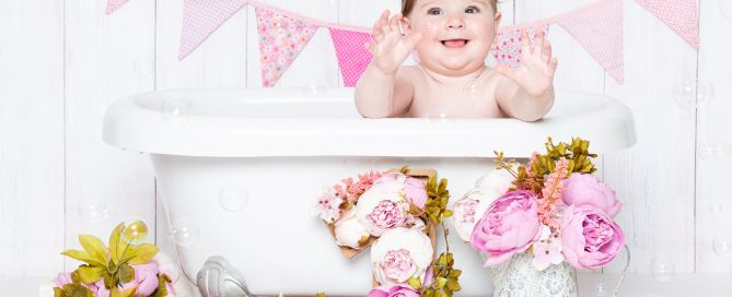 A little girl is excited to be in a bath tub against a white plank backdrop with floral accesories