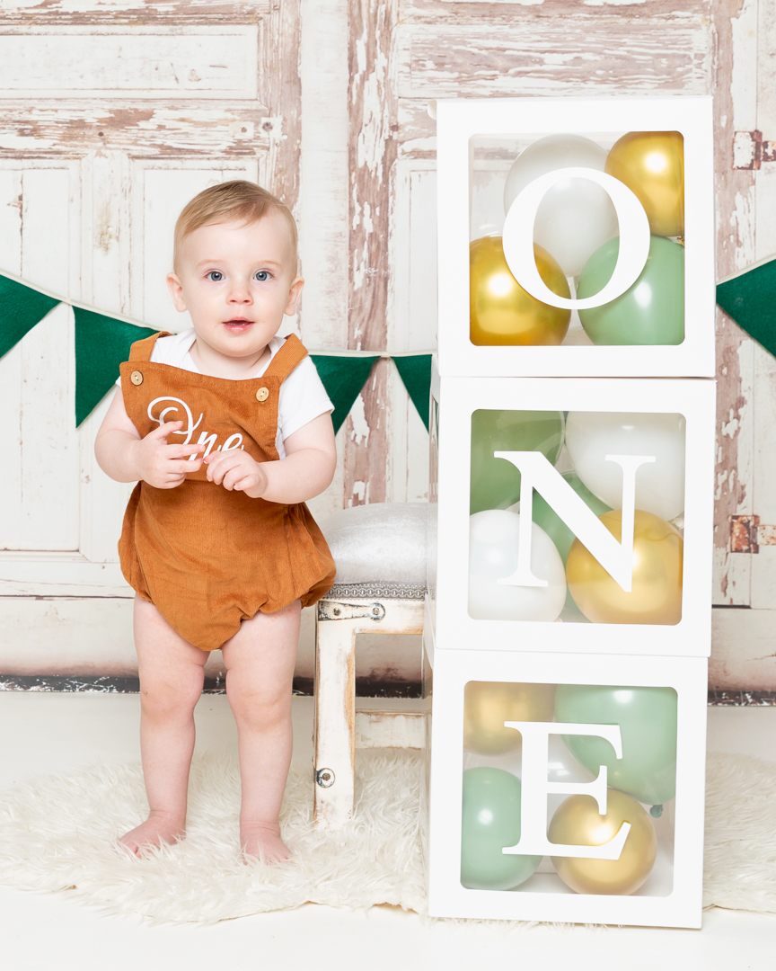 a little boy stands next to a tower of balloon boxes spelling out 'ONE'