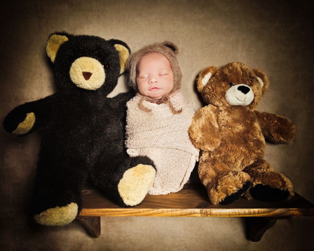 A sleeping newborn in a swaddle and bear hat posed with teddy bears