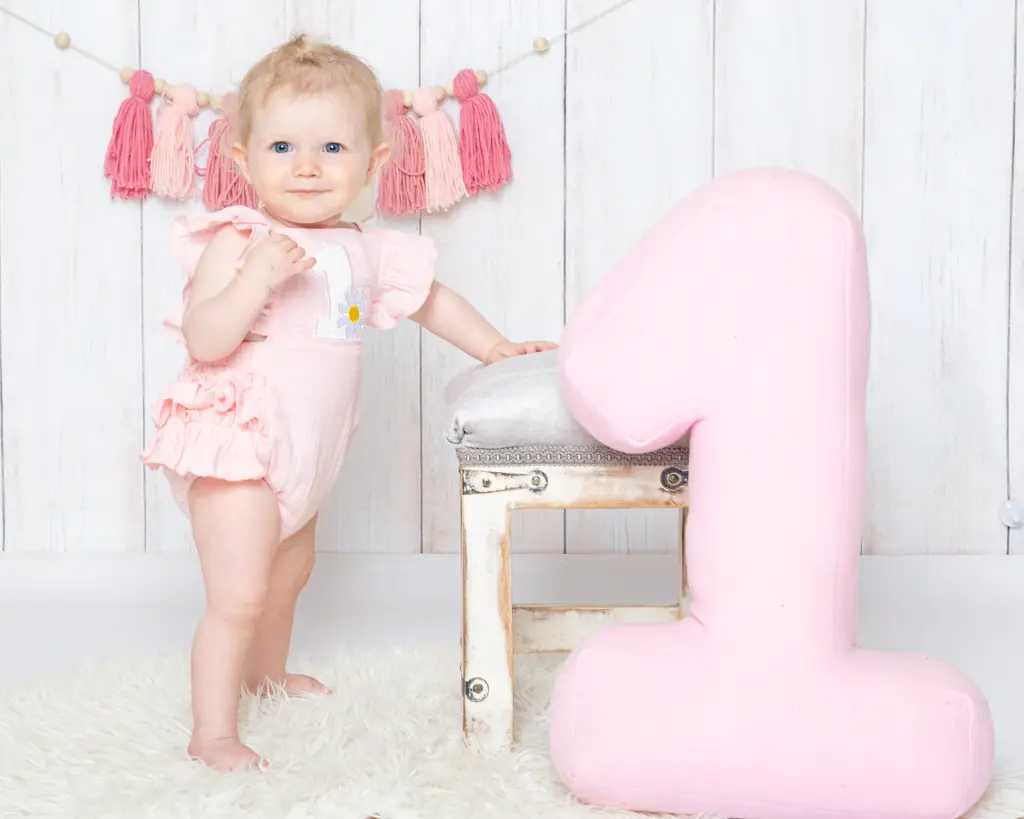 a little girl in a pink romper poses beside a large pink number one at a first birthday photo shoot