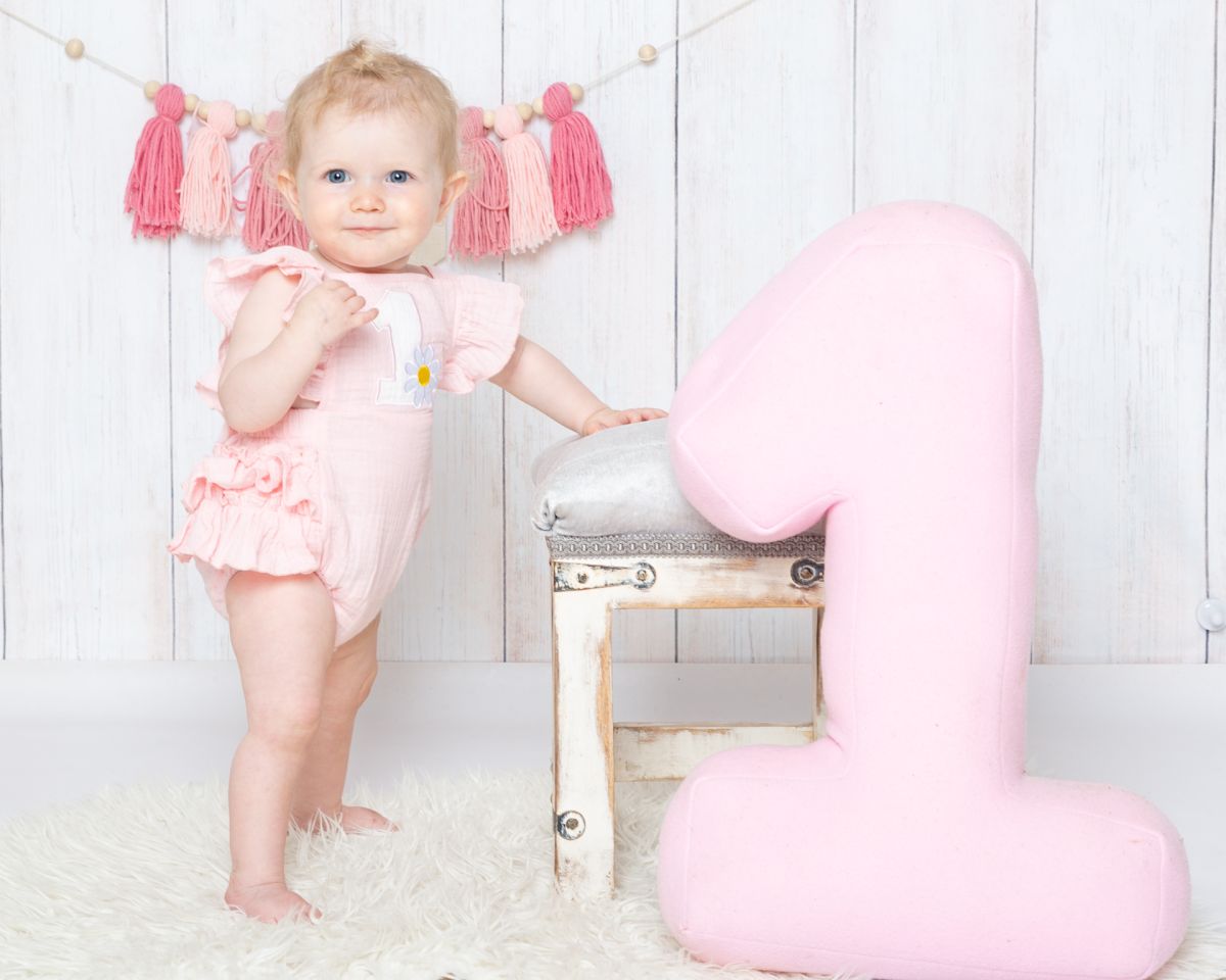 a little girl in a pink romper poses beside a large pink number one at a first birthday photo shoot