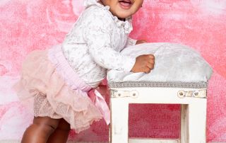 a little girl in a tutu stands up by a footstool against a pink backdrop