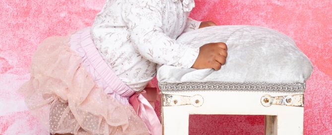 a little girl in a tutu stands up by a footstool against a pink backdrop