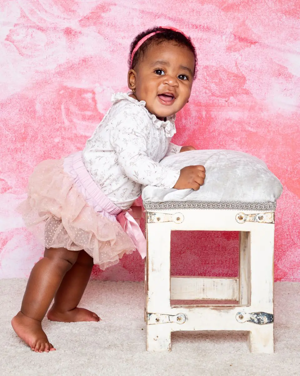 a little girl in a tutu stands up by a footstool against a pink backdrop