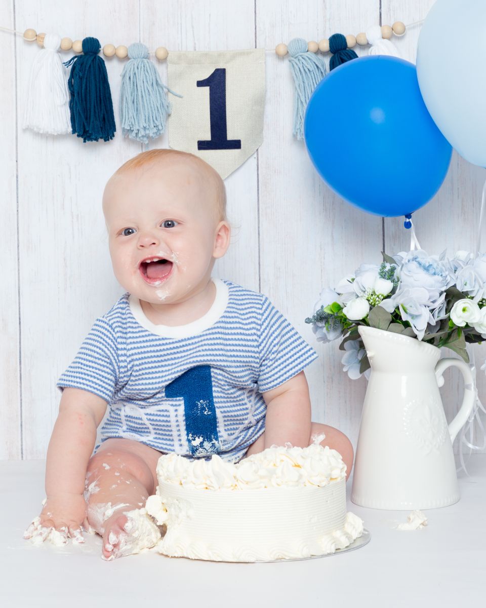 A little boy sits behind a cake with blue balloons and bunting