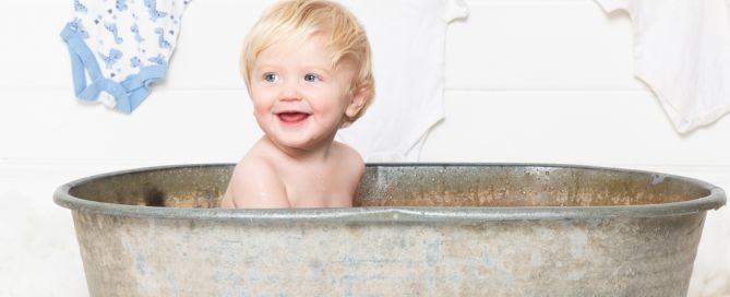 A little boy in a tin tub with laundry hanging on the wall behind him