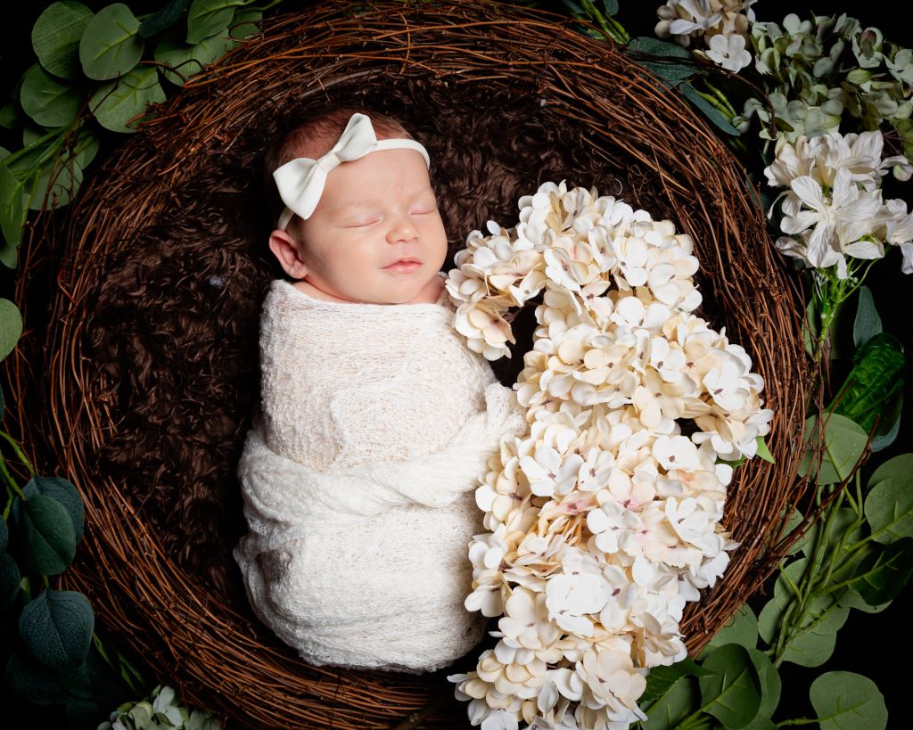 Anewborn is swaddled in white with a matching headband and sleeping in a nest with white flowers around her.