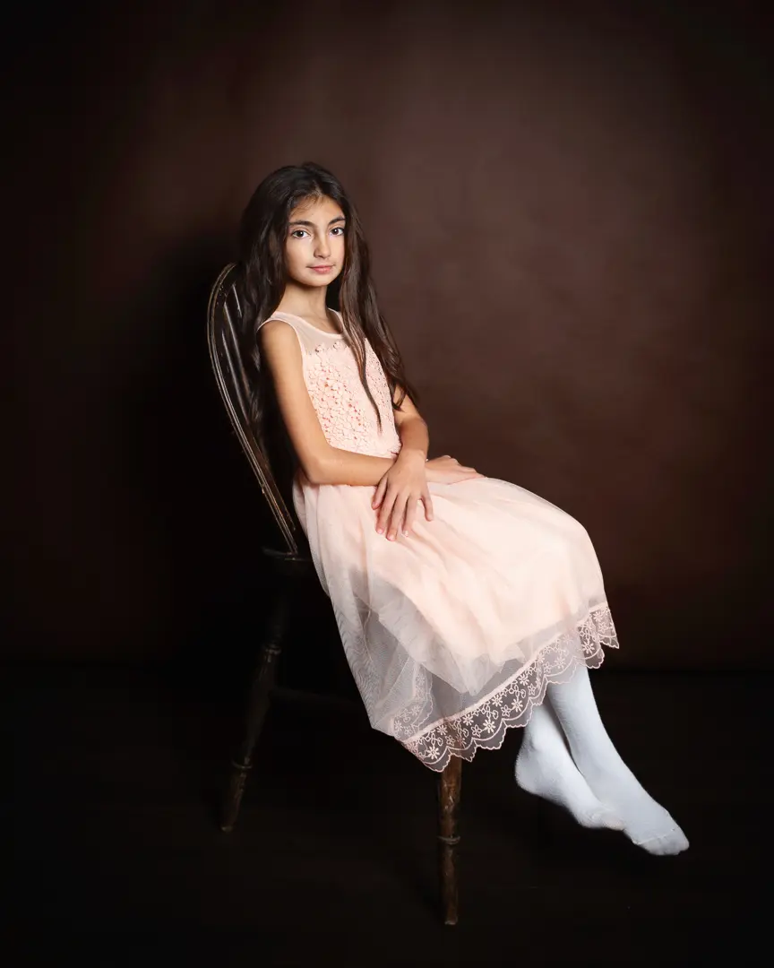 A girl with dark hair poses in a dress on a dark wood chair
