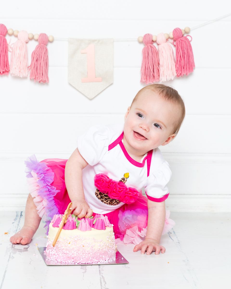 a little girl poses by her cake in a pink pettiskirt with pink tassels behind her - West Sussex