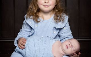 a big sister holds her newborn brother in her lap both dressed in blue