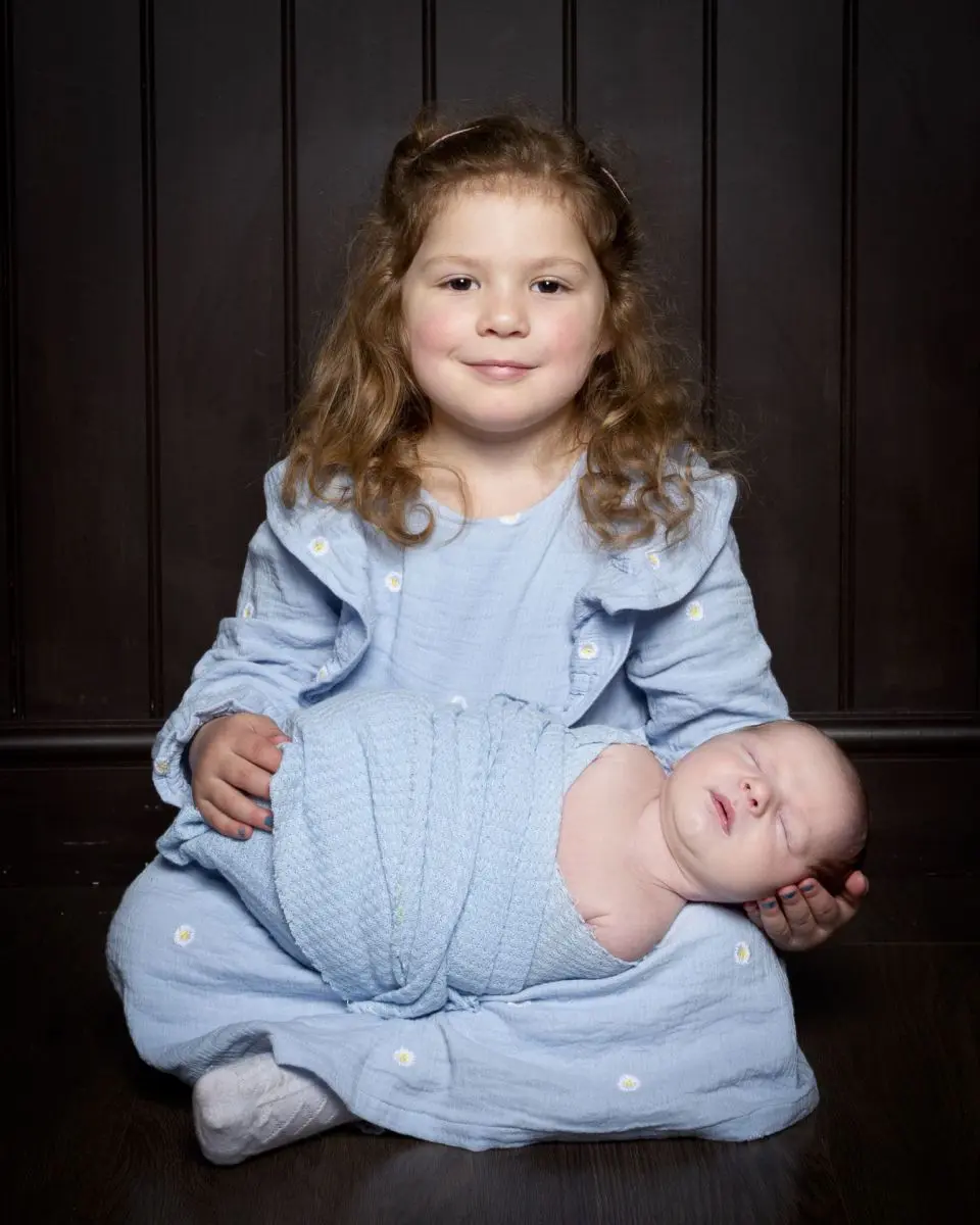 a big sister holds her newborn brother in her lap both dressed in blue