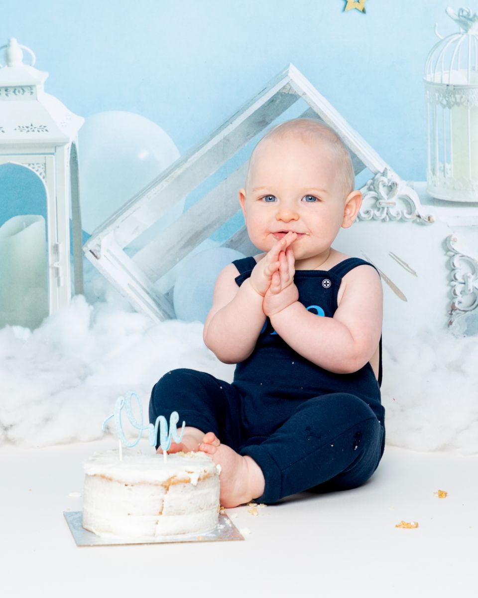 a little boy claps his hands together a she sits behind his cake a first birthday photo shoot.