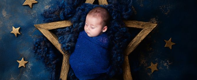 Newborn baby sleeping swaddled in dark blue against a starry sky backdrop