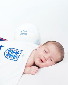 a baby girl sleeps and smiles wrapped in an england football shirt with a football resting behind her.