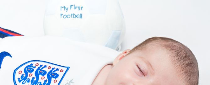 a baby girl sleeps and smiles wrapped in an england football shirt with a football resting behind her.