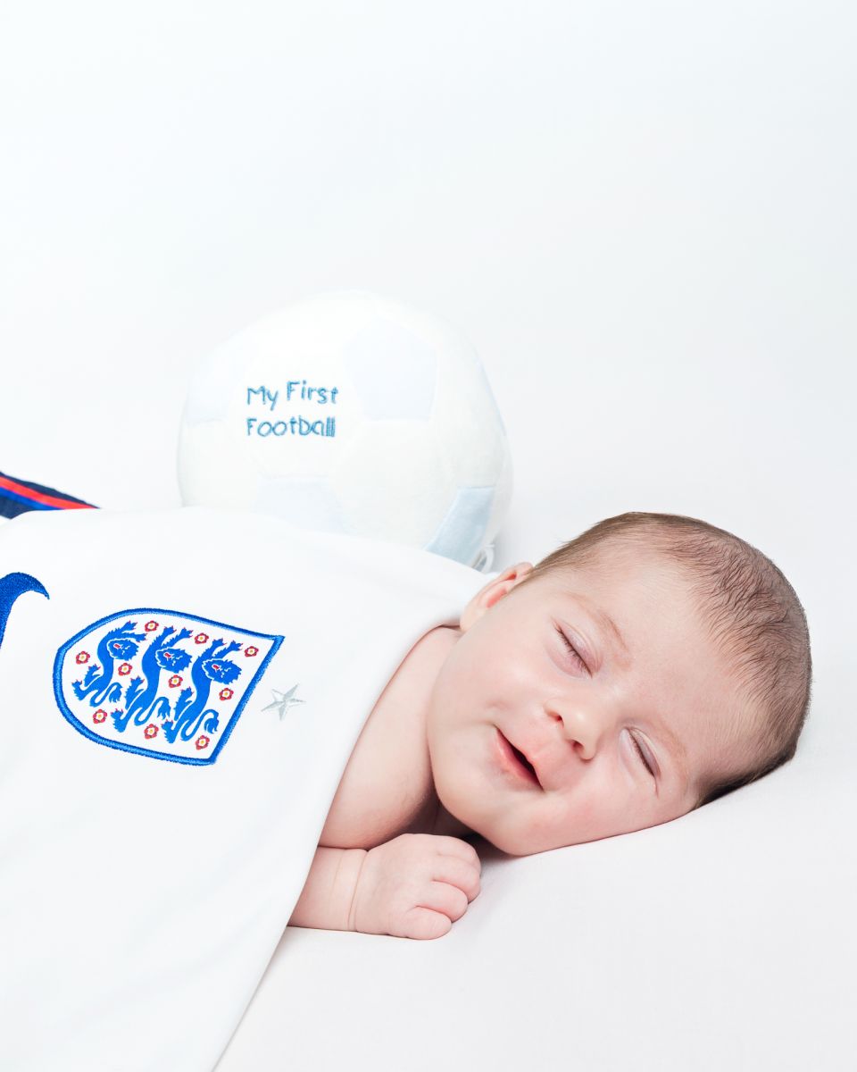 a baby girl sleeps and smiles wrapped in an england football shirt with a football resting behind her.