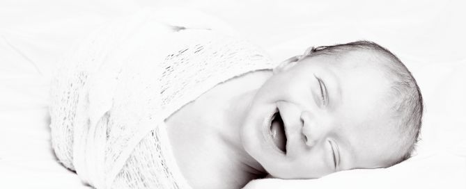 A black and white image of a smiling newborn swaddled in white