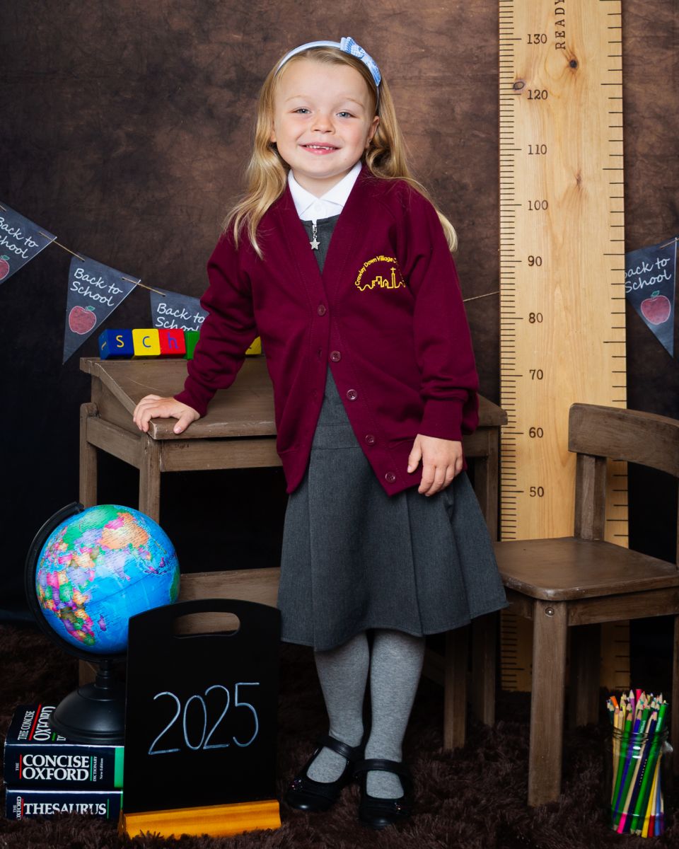A little girl poses by a desk in a burgundy jumper at a Starting School Photo shoot in West Sussex