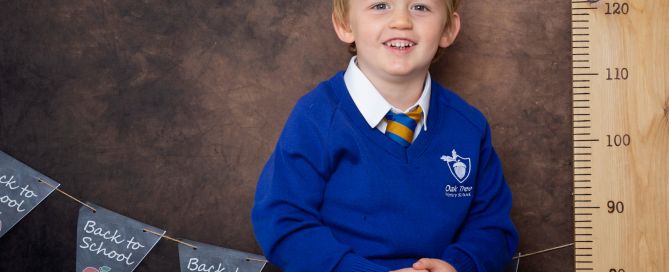 A little boy in a blue jumper poses on a desk at a Starting School Photo shoot in West Sussex