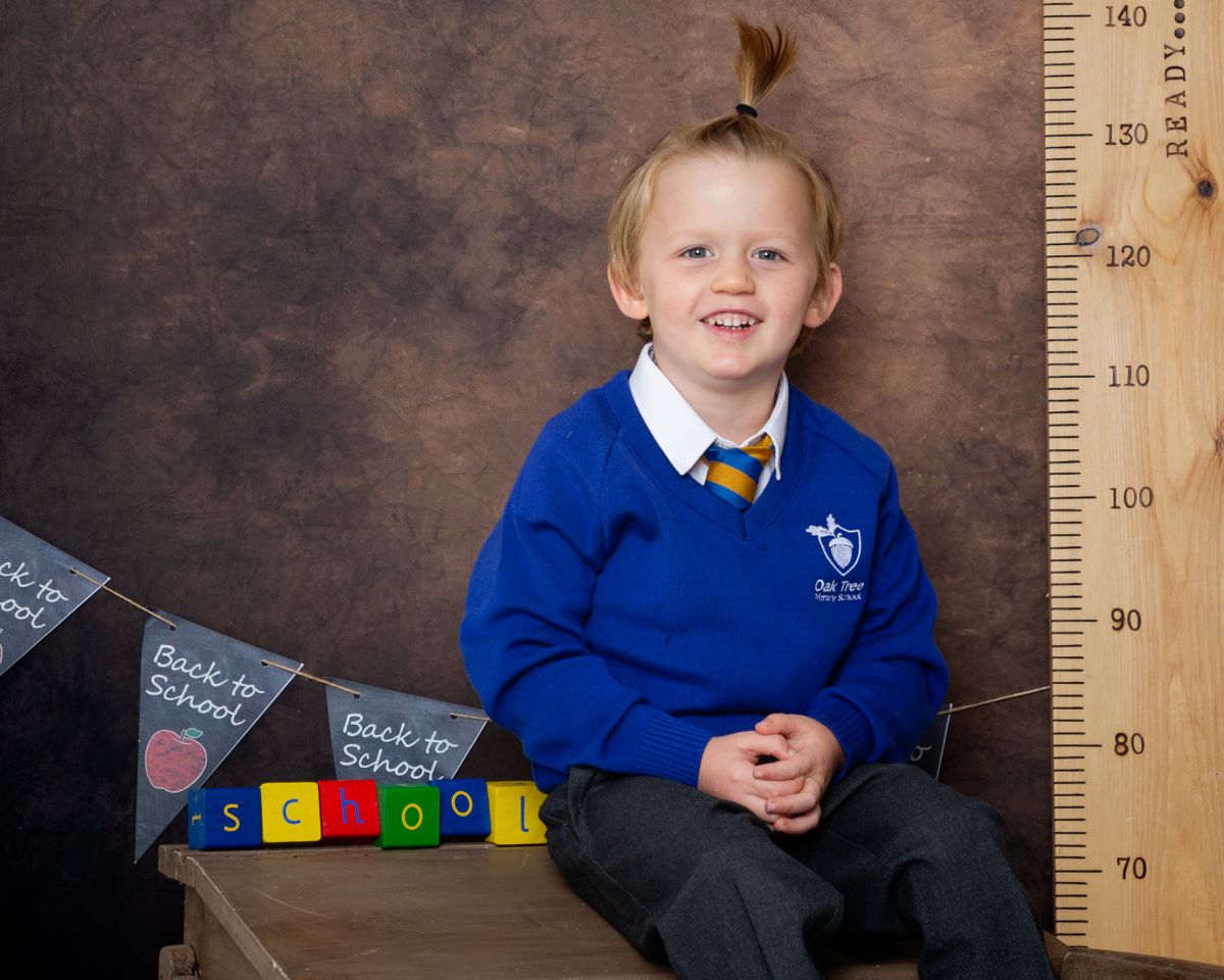 A little boy in a blue jumper poses on a desk at a Starting School Photo shoot in West Sussex