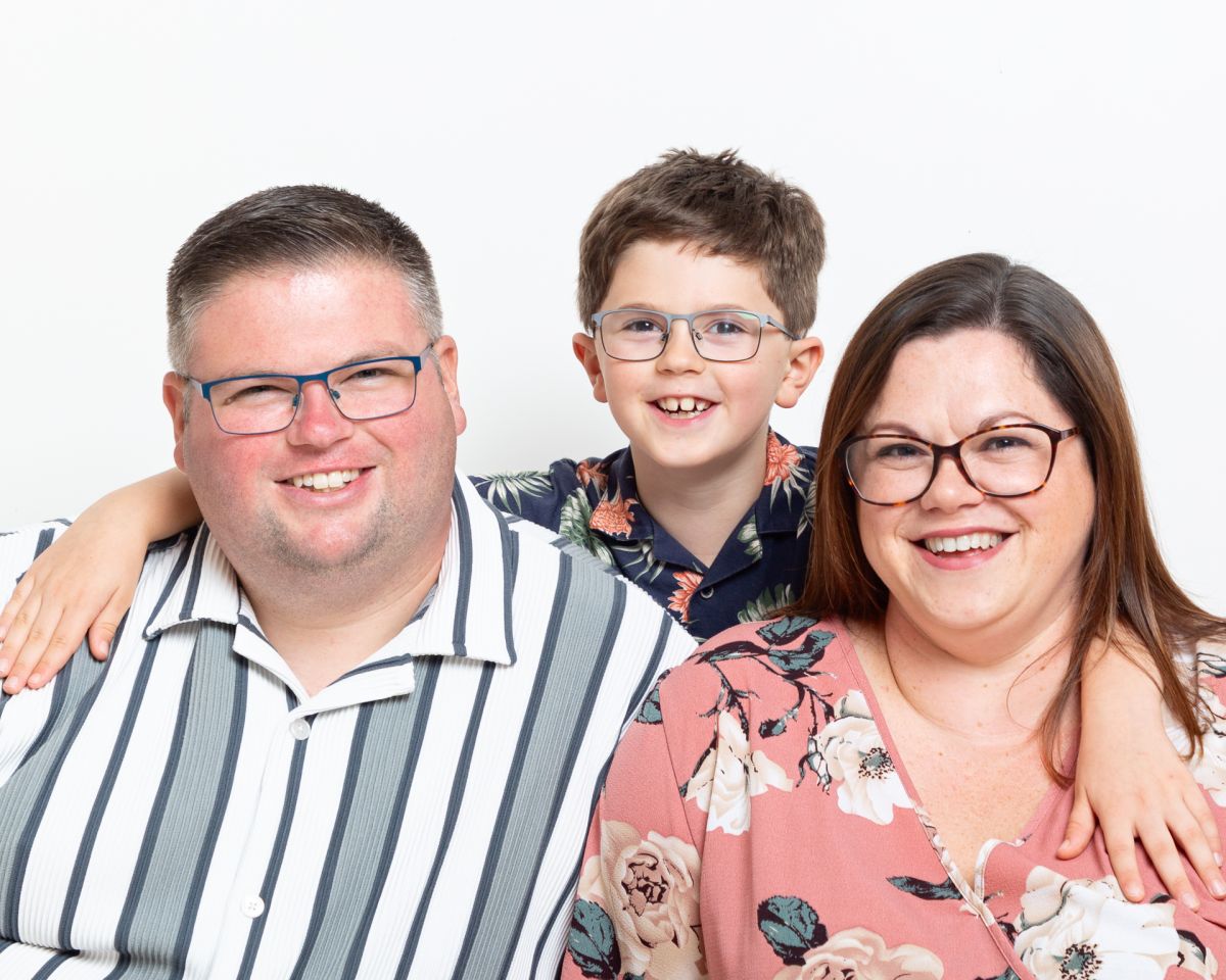 A family of mum, dad and son pose for the camera in a studio against a white background