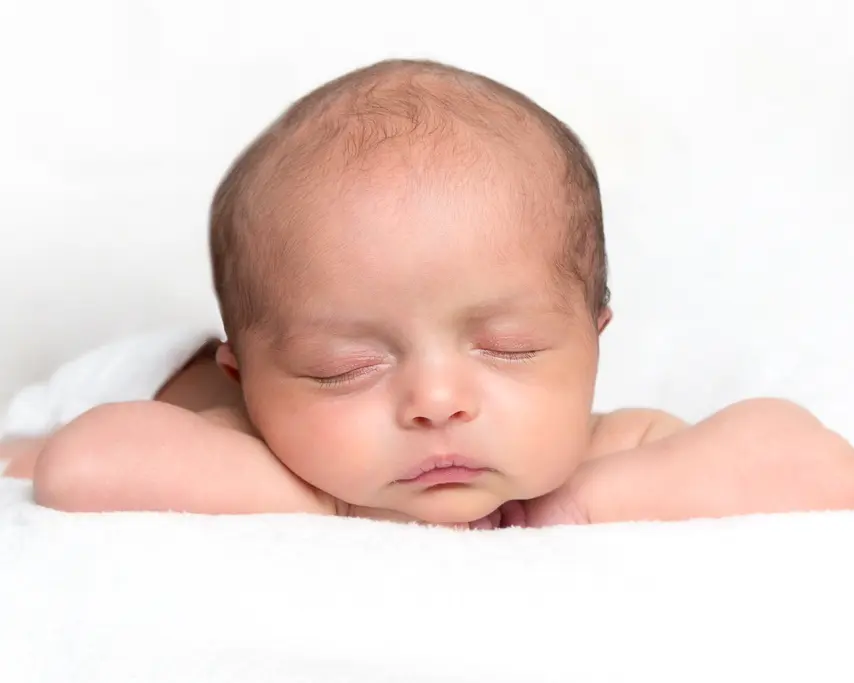 A newborn sleeps on a white blanket with their head on their hands
