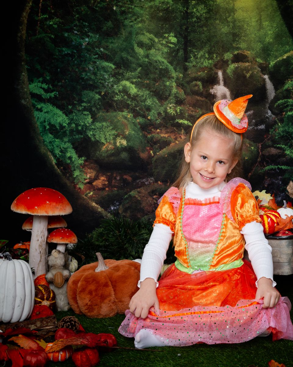 a little girl in an orange witches outfit is sitting in a forest setting