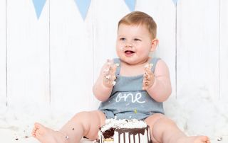 a little boy in a one romper, sits behind his birthday cake