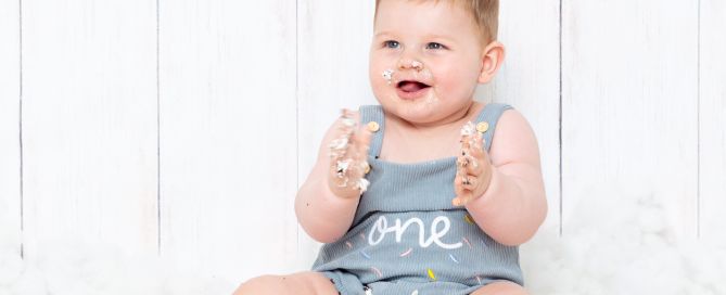 a little boy in a one romper, sits behind his birthday cake