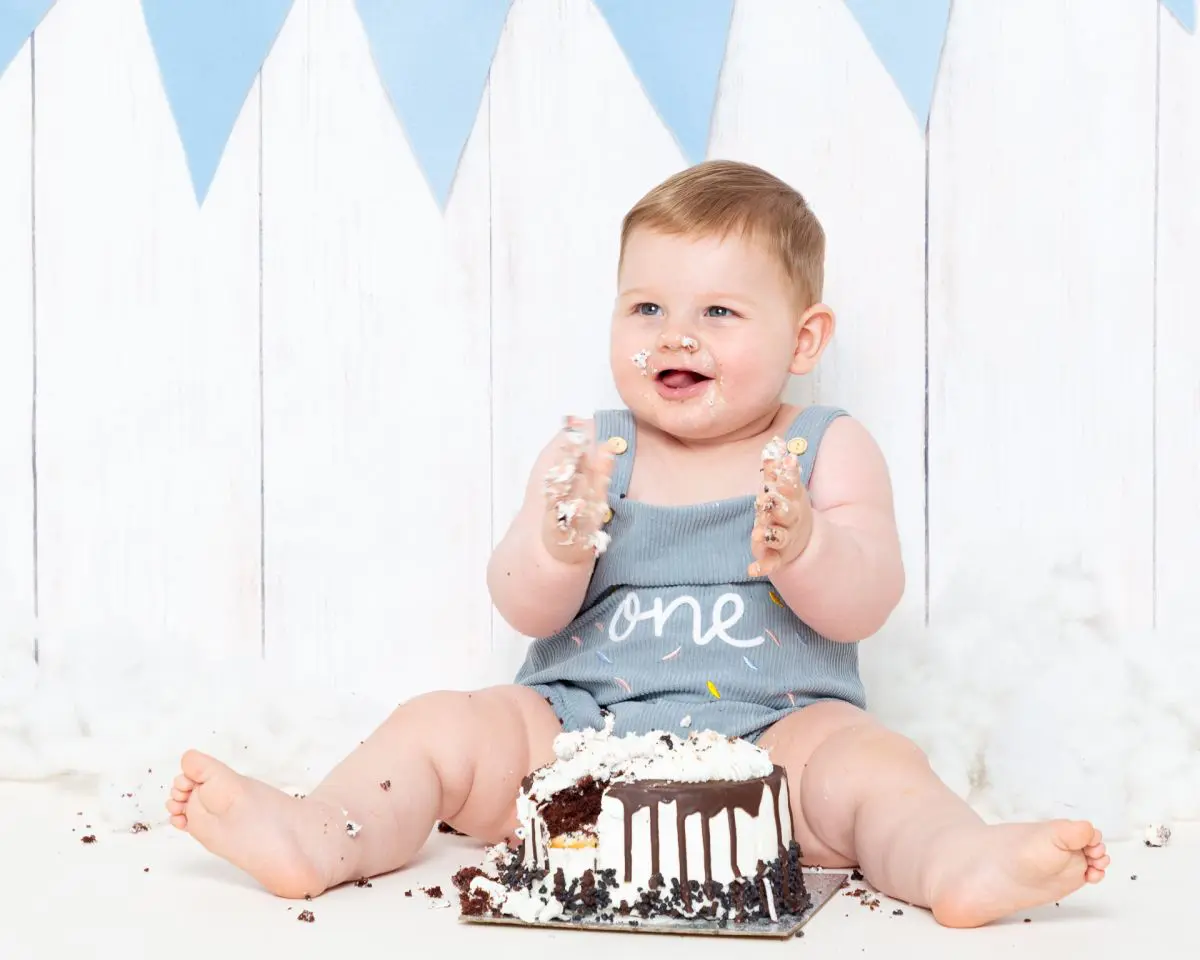 a little boy in a one romper, sits behind his birthday cake