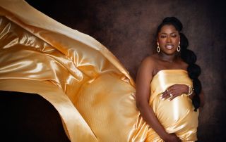 a pregnant woman stands against a deep brown background with a golden fabric flying out behind her